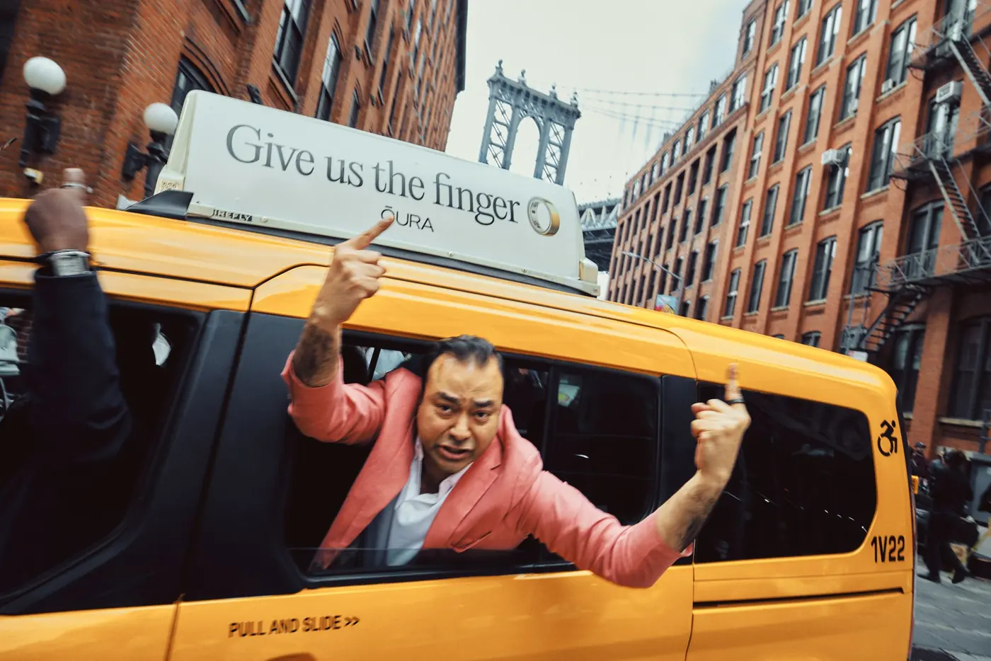 A passenger leans out of a yellow cab under a standard Firefly Icon static top near the Manhattan Bridge