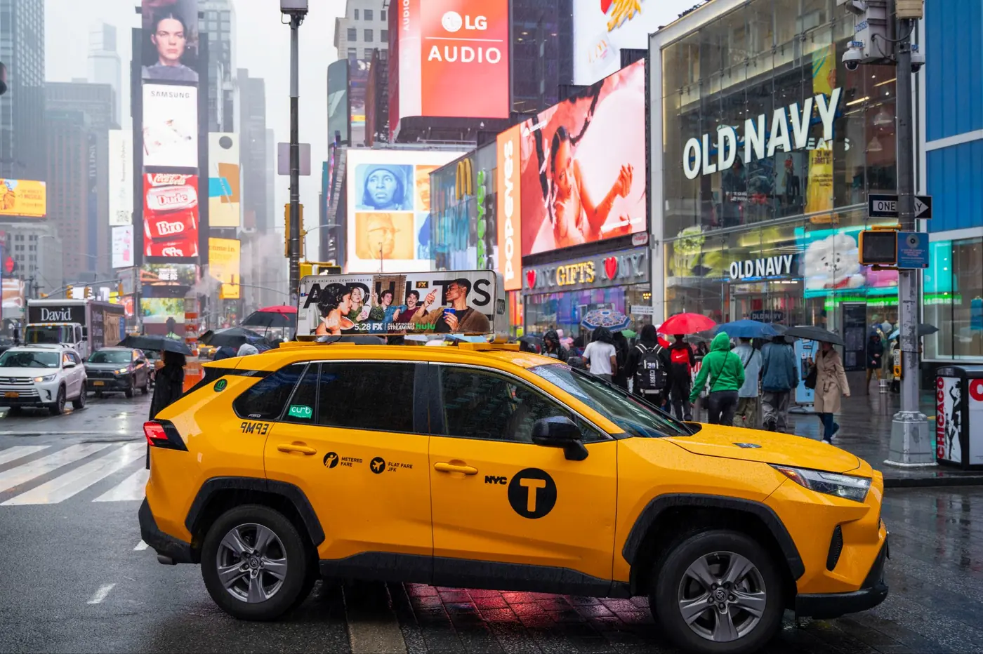 A yellow cab with a static top crossing Times Square past retail storefronts
